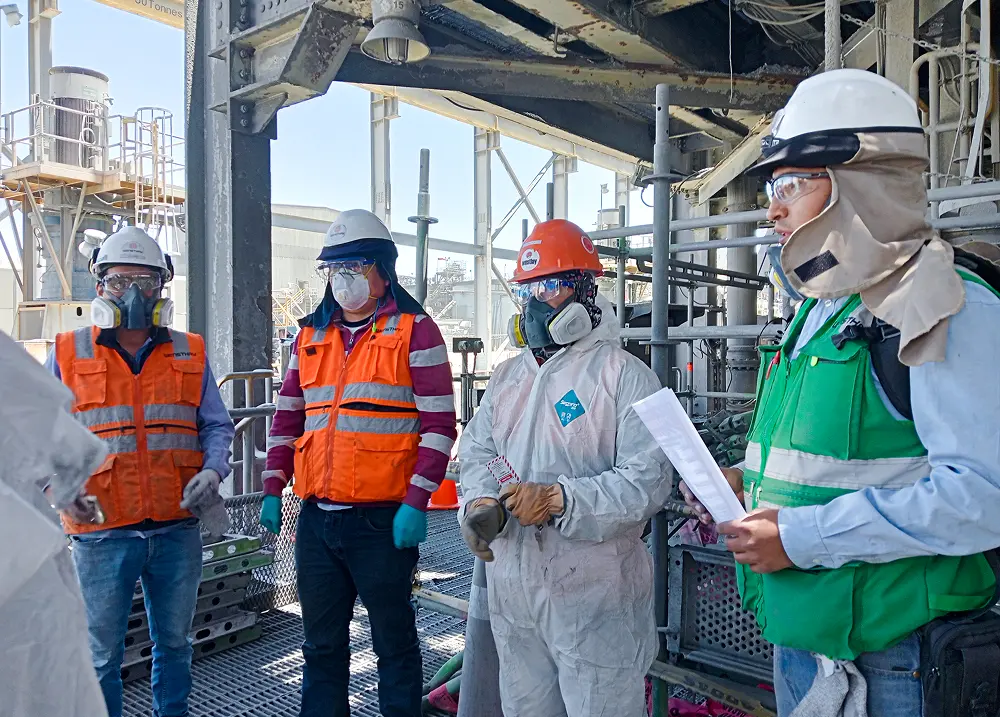 Cuatro trabajadores con equipo de protección, incluyendo cascos y mascarillas, en una instalación industrial.